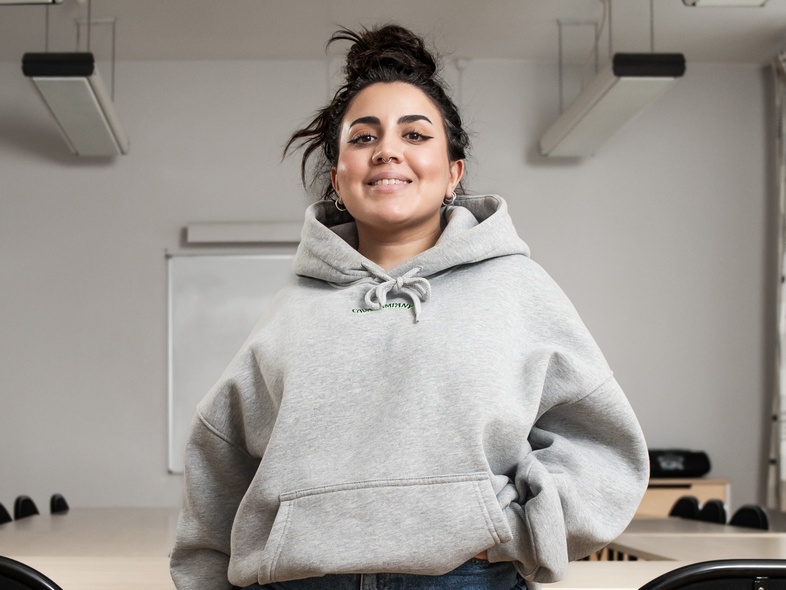 Ung kvinna i grå huvtröja står leende i ett tomt klassrum med bord och stolar.