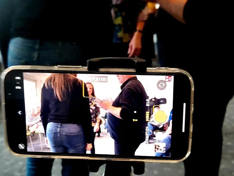 En smartphone på stativ filmar en grupp människor som samtalar inomhus.