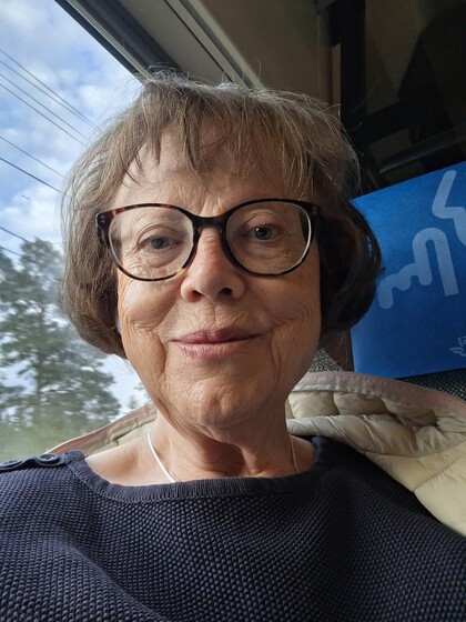 Äldre kvinna med glasögon tar en selfie på ett tåg, med träd och himmel synliga utanför fönstret.