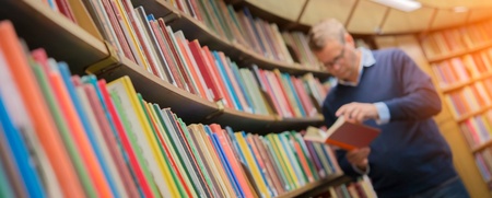 En man står i ett bibliotek och läser i en bok framför en hylla full av färgglada böcker.