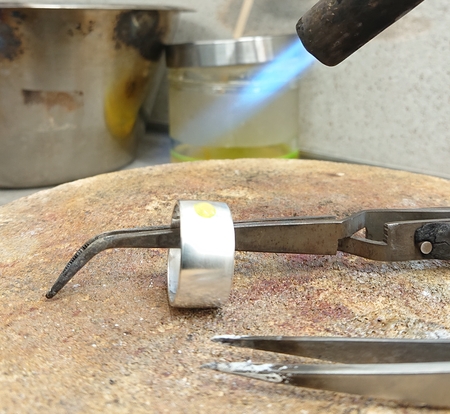 En ring värms upp med en blå gaslåga, hålls med en tång på en arbetsyta i ett smyckesverkstad.