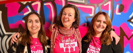 Tre personer står tillsammans framför en färgglad graffitivägg, alla bär rosa t-shirts med texten 'kultur bron'.