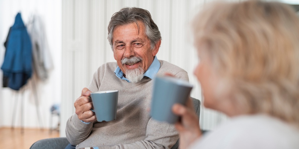 Två äldre personer sitter och dricker kaffe tillsammans i ett vardagsrum.