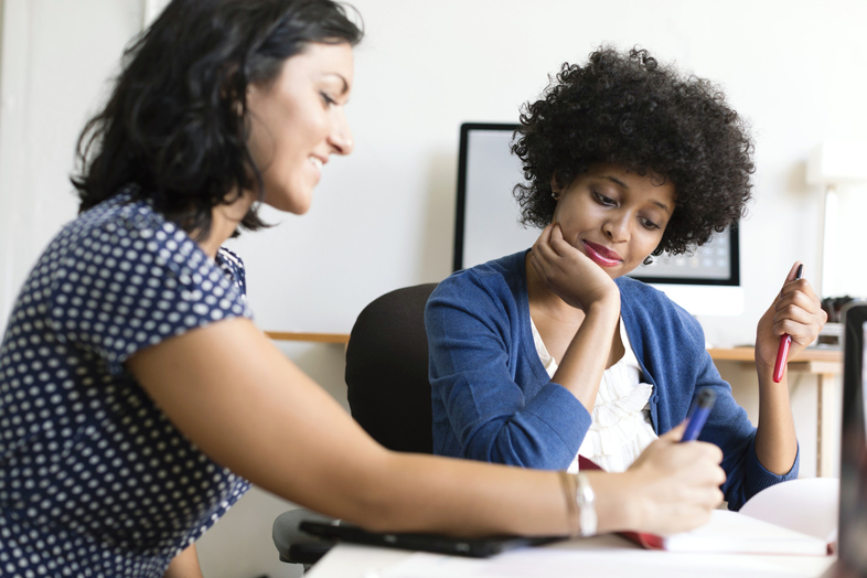 Två kvinnor sitter vid ett bord och arbetar tillsammans med papper och pennor.