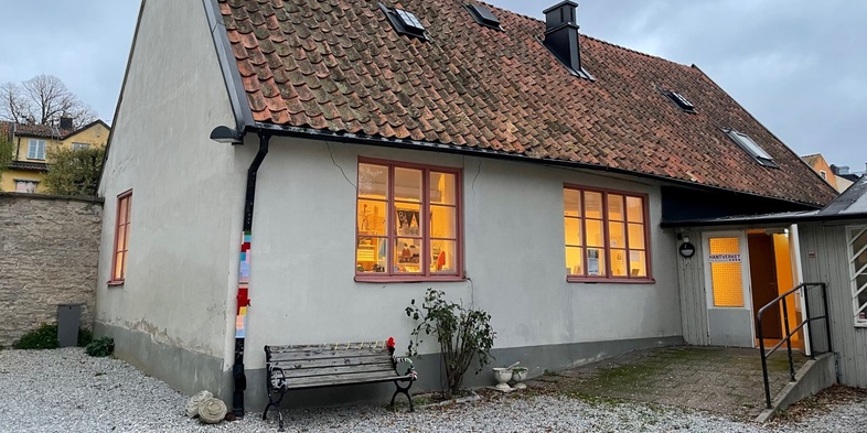 Ett litet hus med tegeltak och grå fasad, upplysta fönster och en bänk utanför, på en grusad gård.