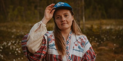 Person i traditionell folkdräkt och blå keps står utomhus i naturmiljö med skog i bakgrunden.
