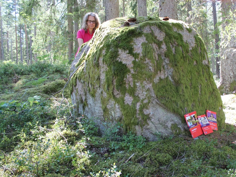 En kvinna står bakom en stor mossig sten i en solig skog. Flera röda broschyrer ligger mot stenen.