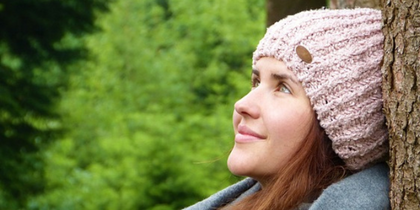 Person med stickad mössa och halsduk lutar mot ett träd i en grön skogsmiljö.
