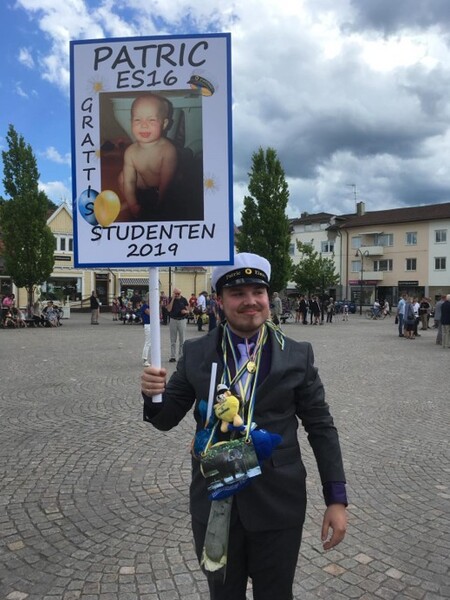 Ung man i kostym och studentmössa håller en skylt med ett barndomsfoto och texten 'Grattis Studenten 2019' på ett torg.