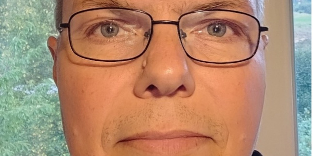 Man med glasögon tittar rakt in i kameran, inomhus med fönster och grönska i bakgrunden.