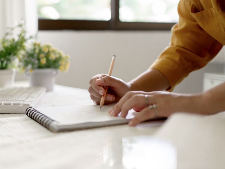 En person skriver i en anteckningsbok vid ett ljust skrivbord med dator, växter och fönster i bakgrunden.