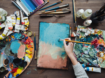En person målar med pensel på en duk omgiven av färgtuber, penslar och paletter på ett träbord.