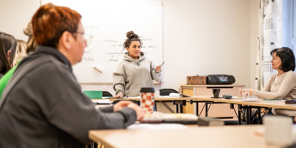 En kvinna står vid en whiteboard och undervisar en grupp vuxna i ett klassrum.