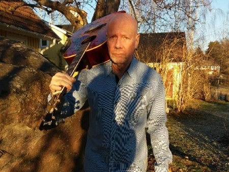 En man står utomhus i solskenet med en gitarr vilande på axeln, omgiven av träd och hus.