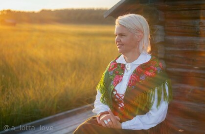 Kvinna i traditionella kläder sitter utomhus vid en timmerstuga i solnedgång.