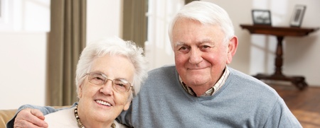 Ett äldre par sitter tillsammans i en soffa och ler mot kameran i ett ljust vardagsrum.