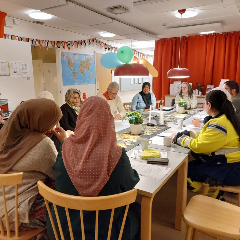 En grupp personer sitter runt ett bord i ett färgglatt rum och har möte eller diskussion.