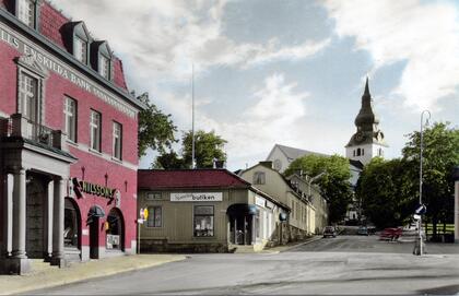 Stadsgata i Hudiksvall med äldre byggnader, bank, butik och kyrka i bakgrunden, några bilar på vägen.