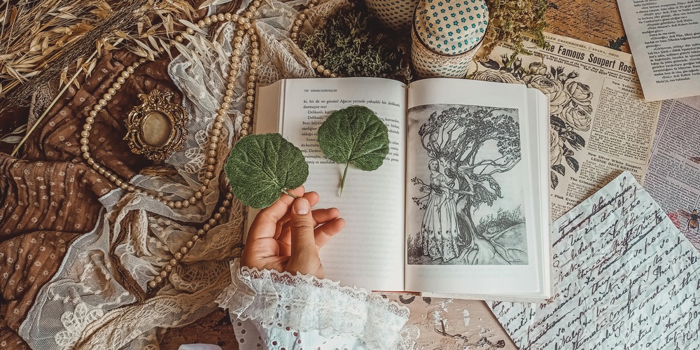 En hand håller två gröna blad över en öppen bok omgiven av vintagepapper, pärlor och spets.