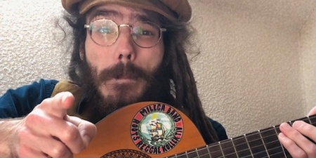 Person sitter och spelar akustisk gitarr dekorerad med klistermärken, iförd basker och bruna byxor.