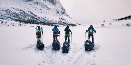 Fyra personer med pulkor står på skidor i ett snötäckt fjällandskap med berg i bakgrunden.