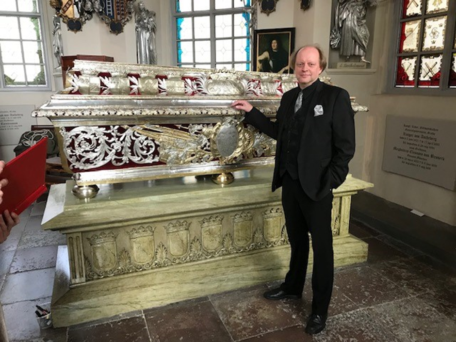 En man i svart kostym står bredvid en utsmyckad sarkofag i ett historiskt rum med färgade fönster.