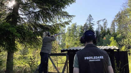 Två personer skjuter lerduvor i en skogsmiljö, en med gevär och en med väst märkt 'Provledare'.