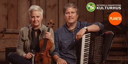 Två personer sitter med musikinstrument, en med fiol och en med dragspel, mot en trävägg. Logotyper syns i hörnet.