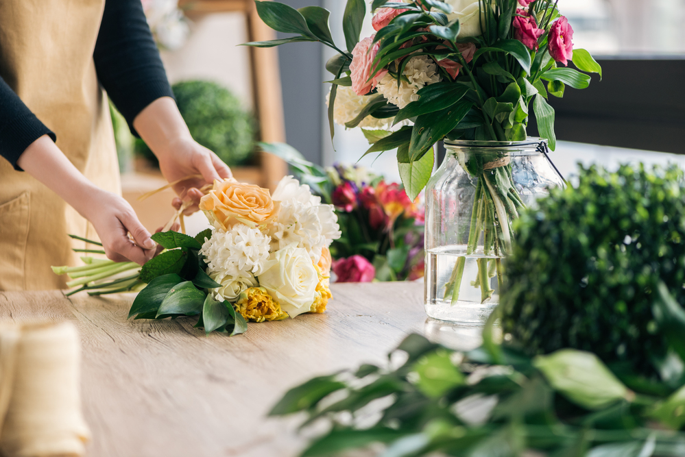 En person arrangerar en bukett med färska blommor på ett träbord bredvid en vas med vatten.