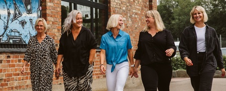 Fem kvinnor går tillsammans utomhus vid en tegelbyggnad med graffiti på väggen.
