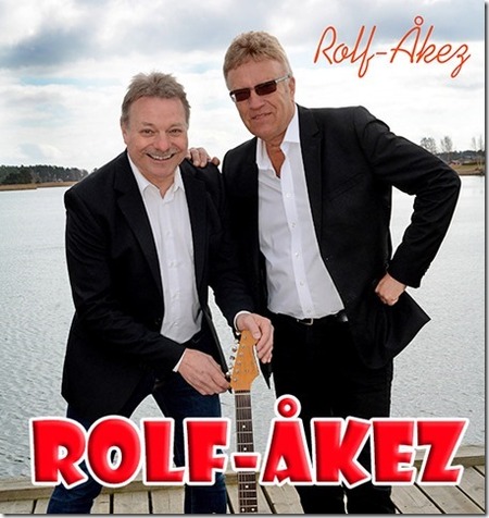 Två män i svarta kostymer står på en brygga vid vattnet, en håller i en gitarr. Texten 'ROLF-ÅKEZ' syns tydligt.