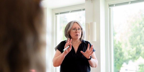 Kvinna med glasögon står och pratar framför ett fönster i ett ljust rum.