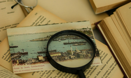 Ett förstoringsglas ligger på en gammal vykort med en hamn, omgiven av öppna böcker.