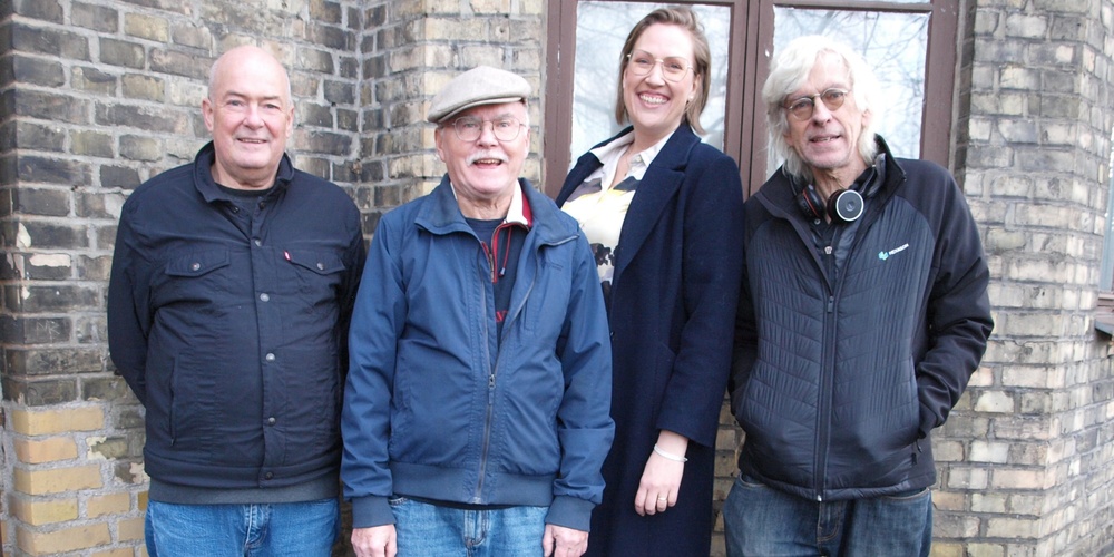 Fyra personer utan instrument står utomhus framför en tegelvägg, tre män och en kvinna.