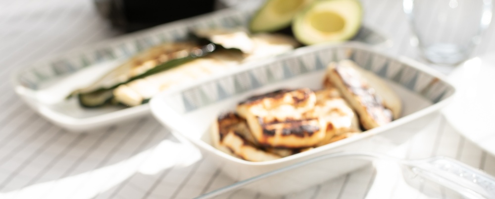 Ett dukat bord med grillad majs, sparris, kyckling, avokado och en vas med lila blommor i solljus.