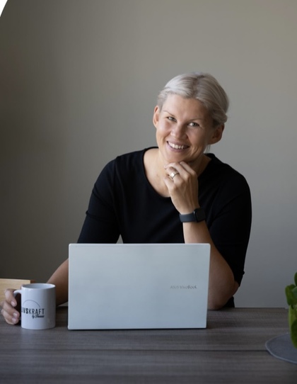 Leende kvinna med ljust hår sitter vid ett bord med en laptop och en kaffemugg.