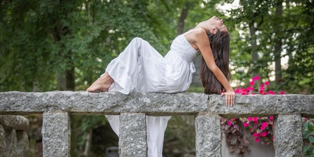 Kvinna i vit klänning poserar liggande på ett stenräcke i en grönskande park.