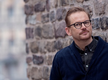 Man med glasögon lutar sig mot en stenmur utomhus, bakgrund med suddiga byggnader.