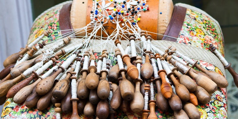 Närbild av en spetskudde med många träspolar och pågående knypplingsarbete.