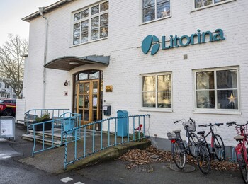 Entré till en vit tegelbyggnad med skylten 'Litorina', cyklar parkerade utanför och bilar på parkeringen.
