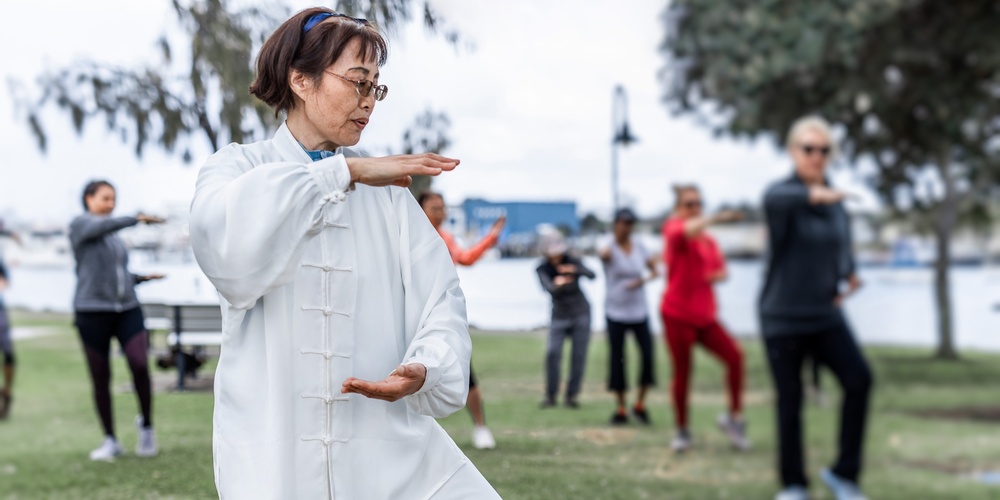 En grupp människor utövar tai chi utomhus i en park, med en kvinna i vit dräkt i förgrunden.