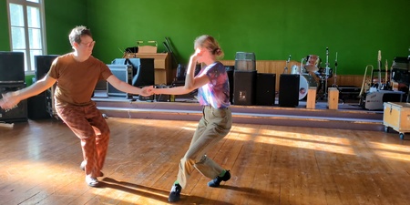 Två personer dansar i ett solbelyst rum med trägolv och musikutrustning i bakgrunden.