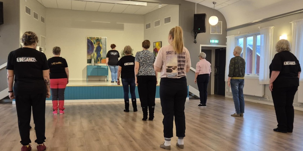 En grupp personer står i ett dansrum och följer en instruktör på en liten scen längst fram.