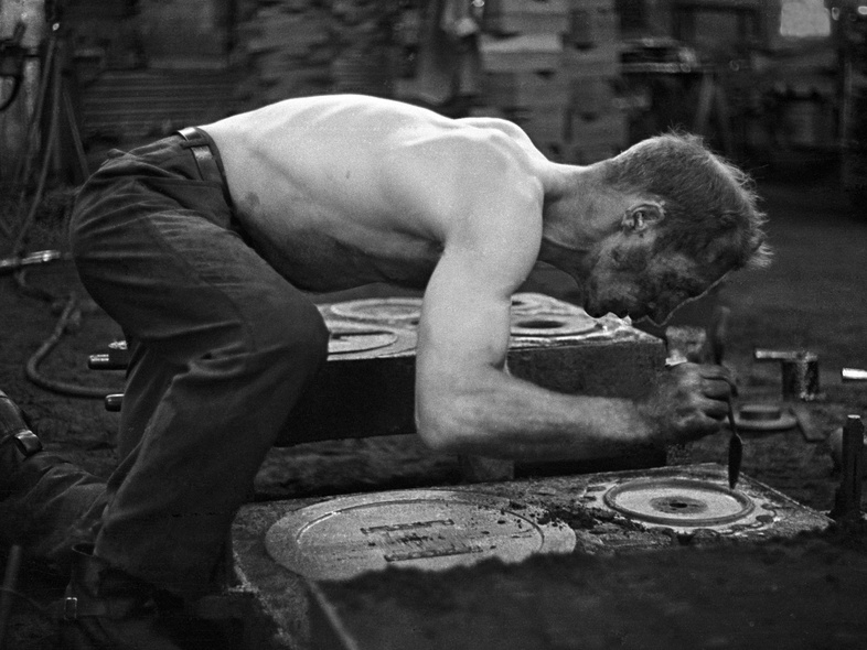 En barbröstad man arbetar böjd över en metallform i en industriell verkstadsmiljö.