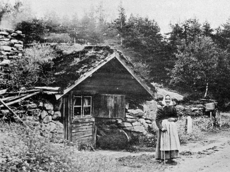 En kvinna i traditionella kläder står framför ett litet torp med grästak vid en landsväg, omgiven av skog.