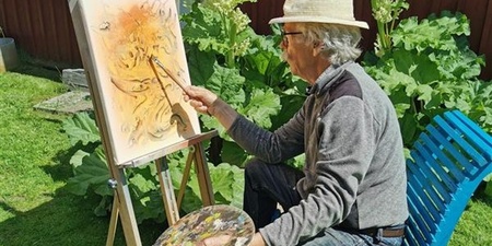 En person målar på en duk utomhus, sittande på en blå stol med en palett i handen.