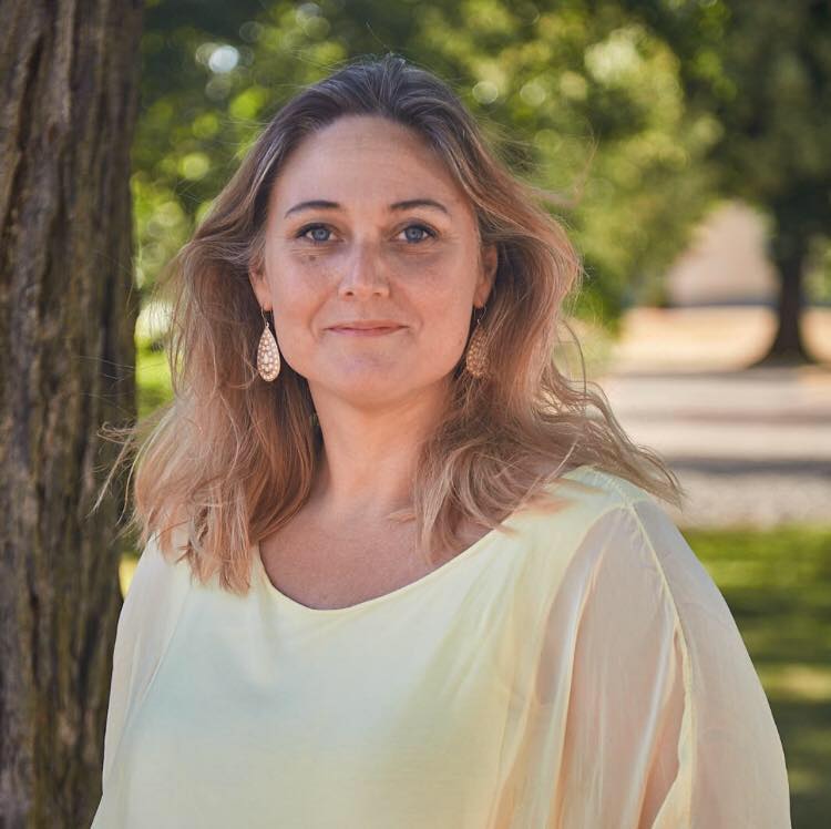 Kvinna med ljust hår i ljusgul blus står utomhus vid ett träd, med grönskande bakgrund.