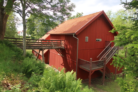 Röd träbyggnad med utvändig trappa och gångbro, omgiven av grönska och träd.
