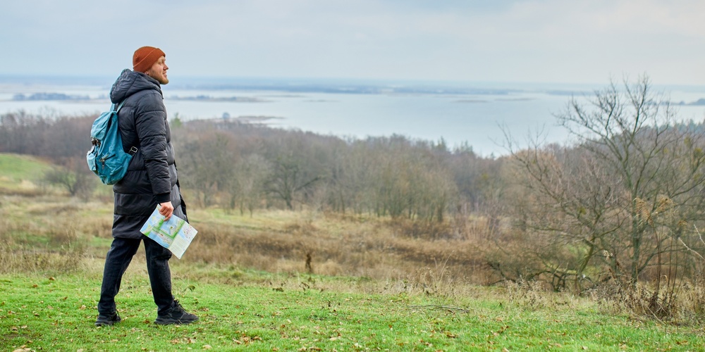 En person med ryggsäck och karta står på en grön äng med utsikt över sjö och skog.