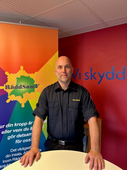 Man i mörk uniform står vid ett bord framför en färgglad informationsskylt och en röd vägg med blå text.